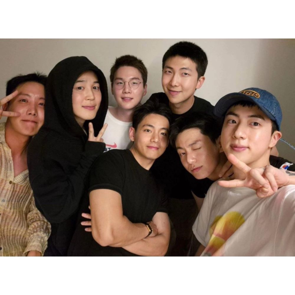 BTS members pose together indoors for a close-up group selfie, all smiling or making playful expressions. They are dressed casually, with some wearing t-shirts and Jiminie in a hoodie.