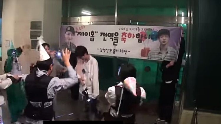 Seokjin, Hobi, and zombies celebrating outside of an abandoned hospital, with party hats, cake and a banner.