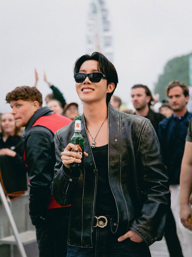 Hobi standing outside and watching the Lollapalooza stage in a barricaded off spot. He's wearing sunglasses, a black leather jacket, a black tanktop underneath.