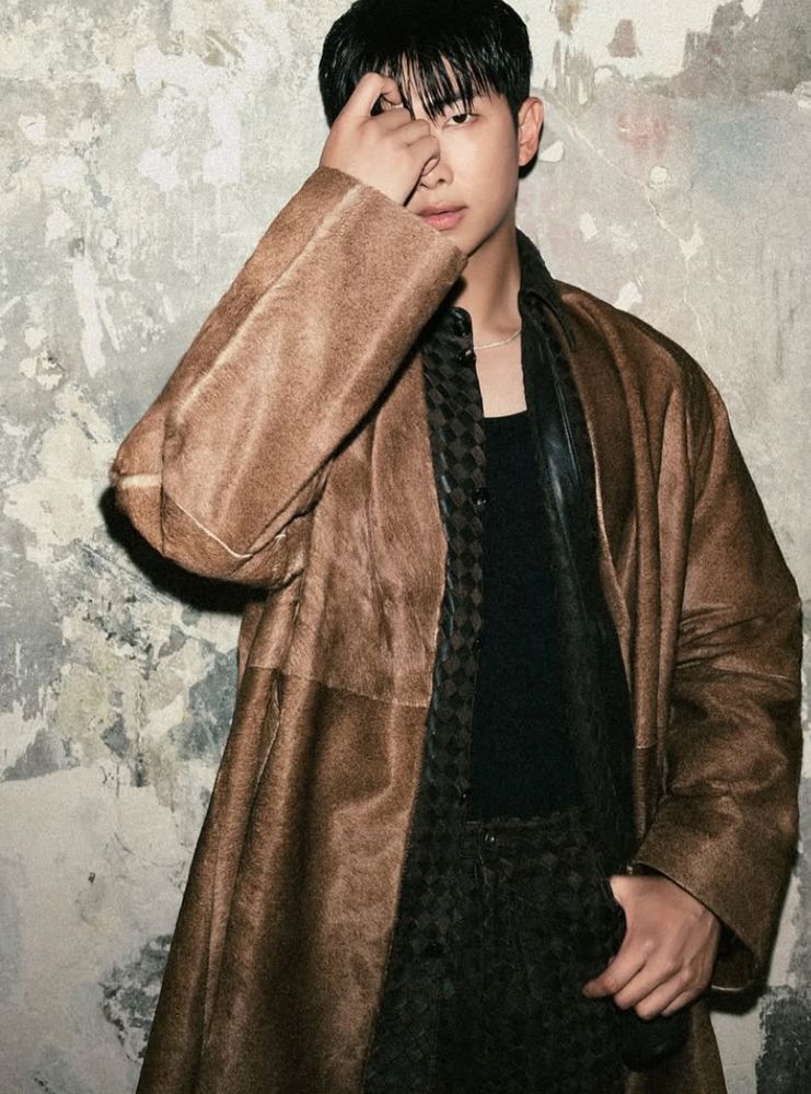 Namjoon in black and brown velvet-looking material / robe, standing in front of a textured wall. His face partly covered by his hand. 