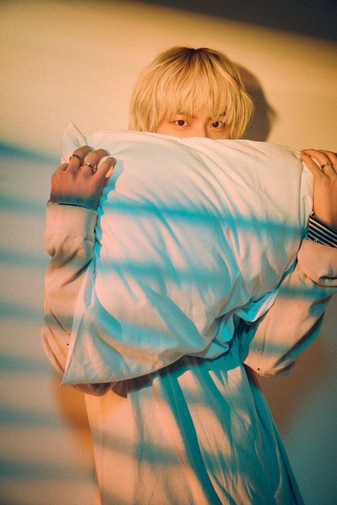 Blonde seokjin standing against the wall hugging a pillow. A yellow light filtering through from the blinds. 
