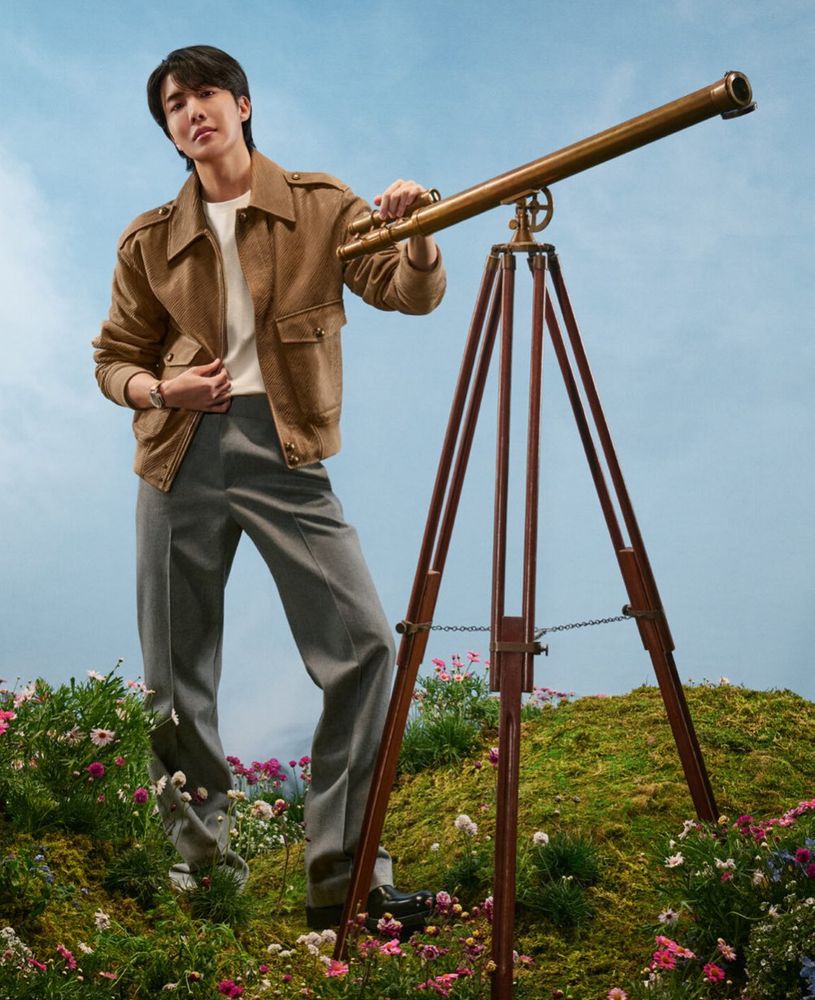 J-hope in gray slacks and brown jacket over white top standing by a telescope on an outdoor set