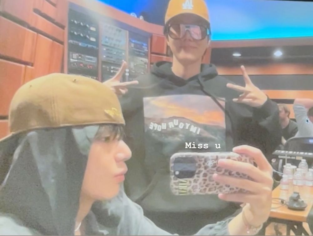 Taehyung taking a mirror selfie of him and Hobi, both dressed casually hanging out in what looks like a studio. Other people can be seen in the background. 
