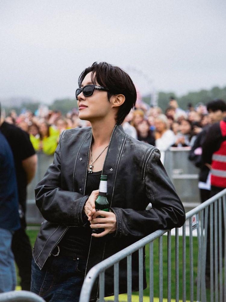Hobi standing outside and watching the Lollapalooza stage in a barricaded off spot. He's wearing sunglasses, a black leather jacket, a black tanktop underneath.