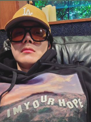 Hobi selfie. He's wearing a hoodie with a fake Hollywood sign that says I'm Your Hope. He's also wearing sunnies and a yellow cap.
