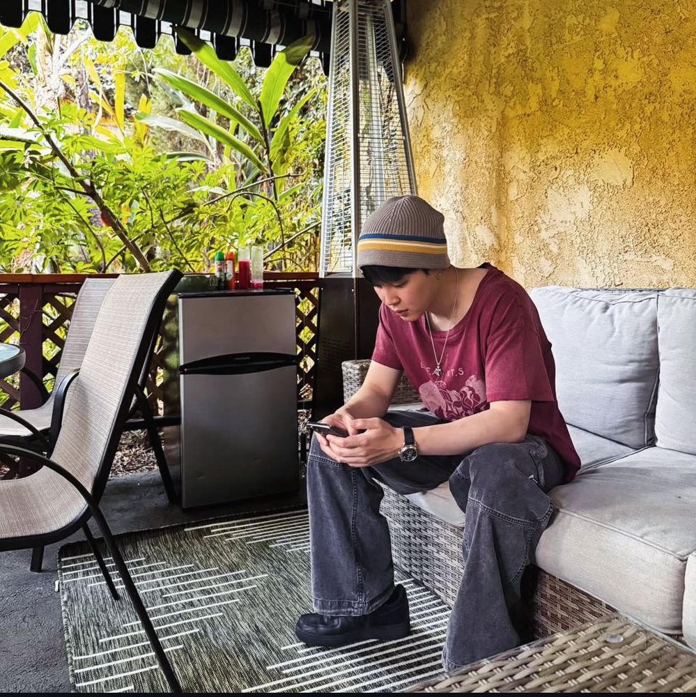 Jimin sitting outdoors on a a big sofa on the patio dressed casually in a printed tee and jeans and a beanie. He’s focused on his phone. 