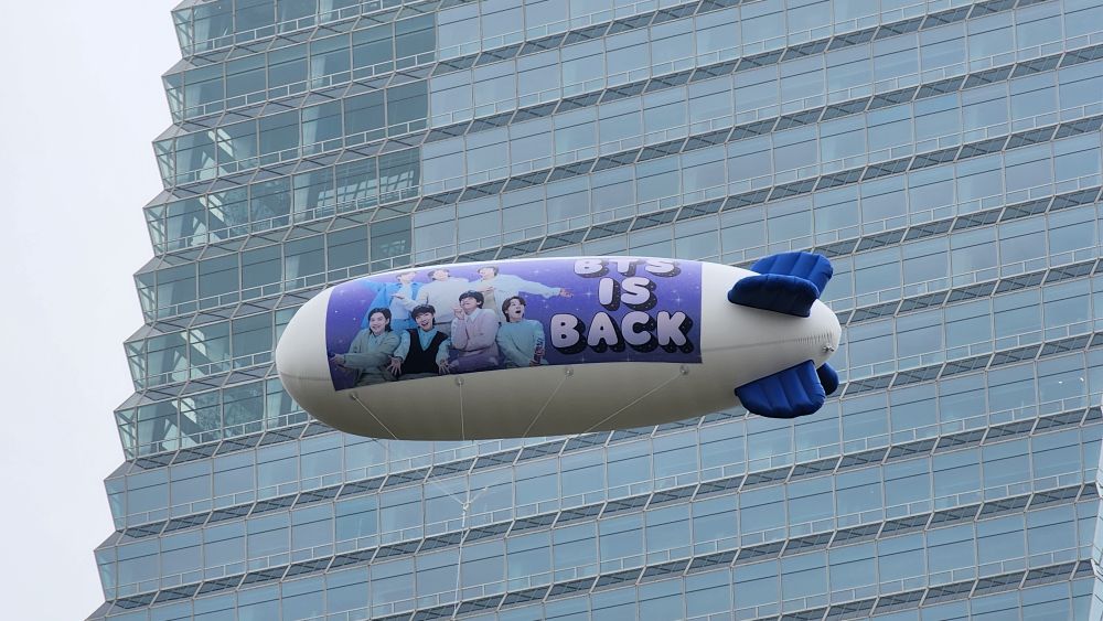 A large white and blue blimp floats in the sky near a glass high-rise building. The blimp features a vibrant banner with all seven BTS members smiling and posing against a starry purple background. Bold text on the banner reads “BTS IS BACK.”