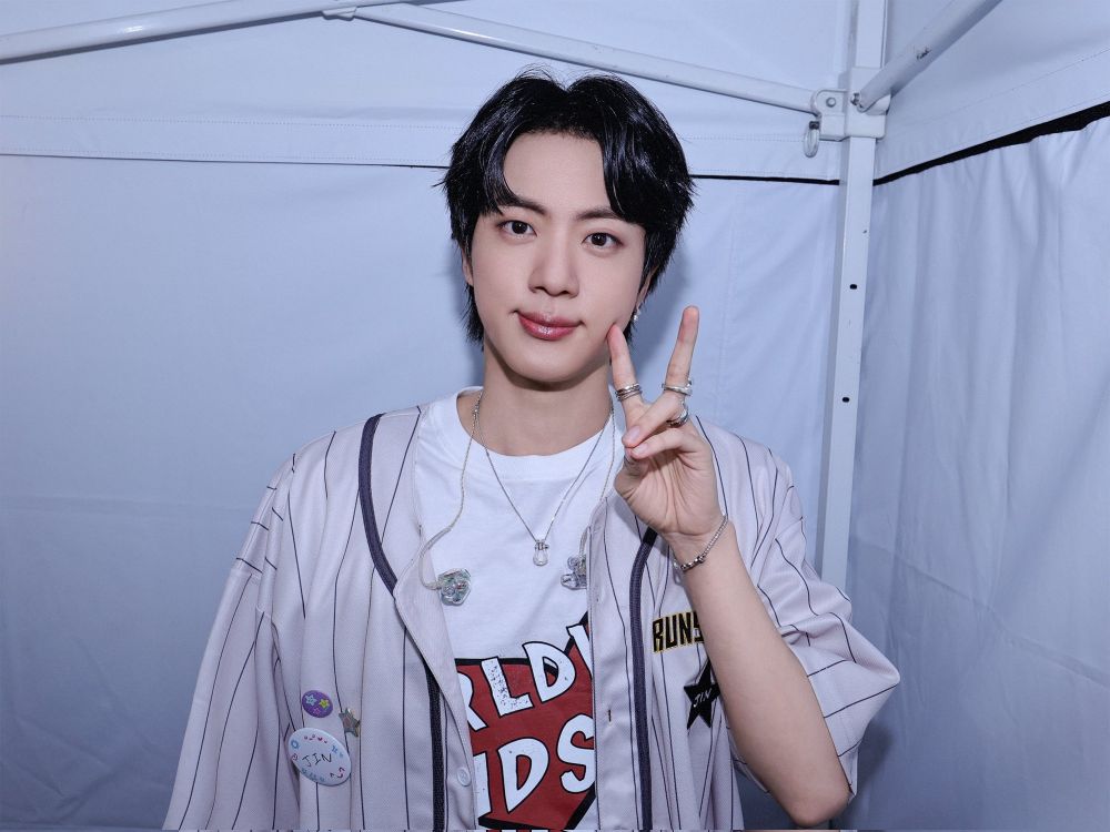 Seokjin in his baseball jersey merch, making s peace sign