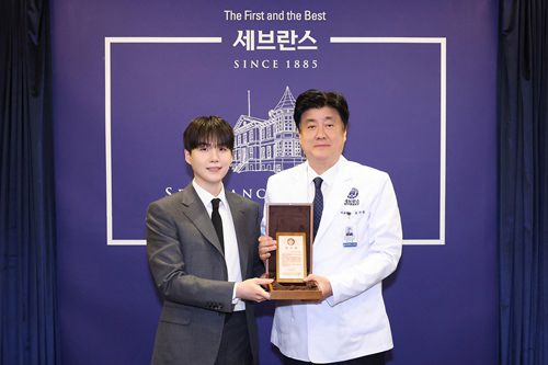 Yoongi dressed in a dark gray suit and black tie, stands beside a doctor in a white medical coat. They are both holding a wooden plaque or award together and posing for a photo in front of a dark blue backdrop with the Severance Hospital logo and the text “The First and the Best, Severance, Since 1885.”