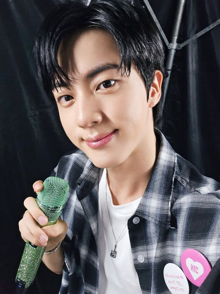 Seokjin in plaid flannel shirt over white inner shirt, taking a selfie holding his green mic