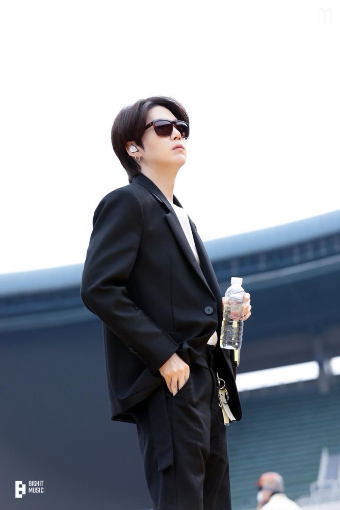 Yoongi standing on an outdoor stage holding a water bottle and a microphone in one hand, while his free hand his reaching in his pocket. He's wearing sunglasses, a black jacket, a white shirt underneath, and dark pants. 