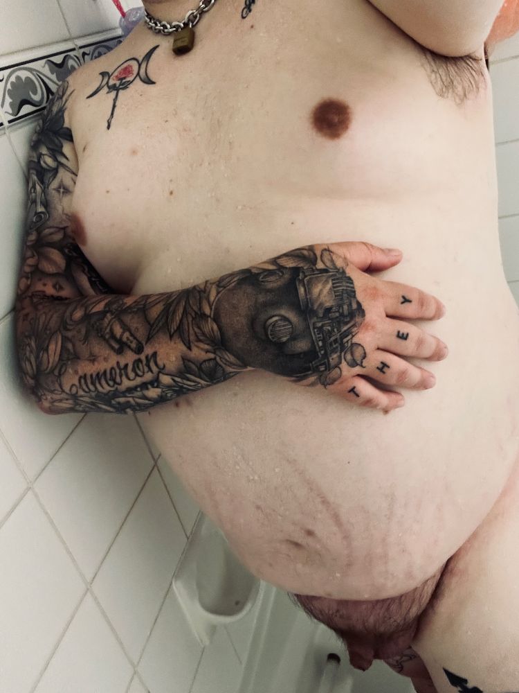 a selfie of naked creature, from its neck down to its thighs, standing in its white tiled shower. it’s right tattooed arm is across its chest under its boobs while the left arm is outstretched holding the camera. its chain collar and goddess rose chest tattoo are visible at the top of the frame. at the bottom of the frame alone the left side is the wall of the shower that creature is leaning on. it’s right leg is in the lower right corner of the frame. only the top of the thigh and a portion of the trans symbol tattoo is visible. creatures genitals are also visible
