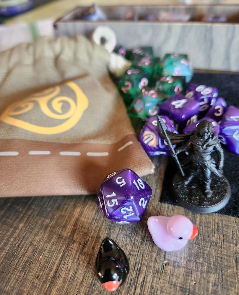A collection of purple and green dice with a dice bag styled as a heavy sack from ESO. A character mini holding a spear is also there, along with two ducks, one black and one pink.