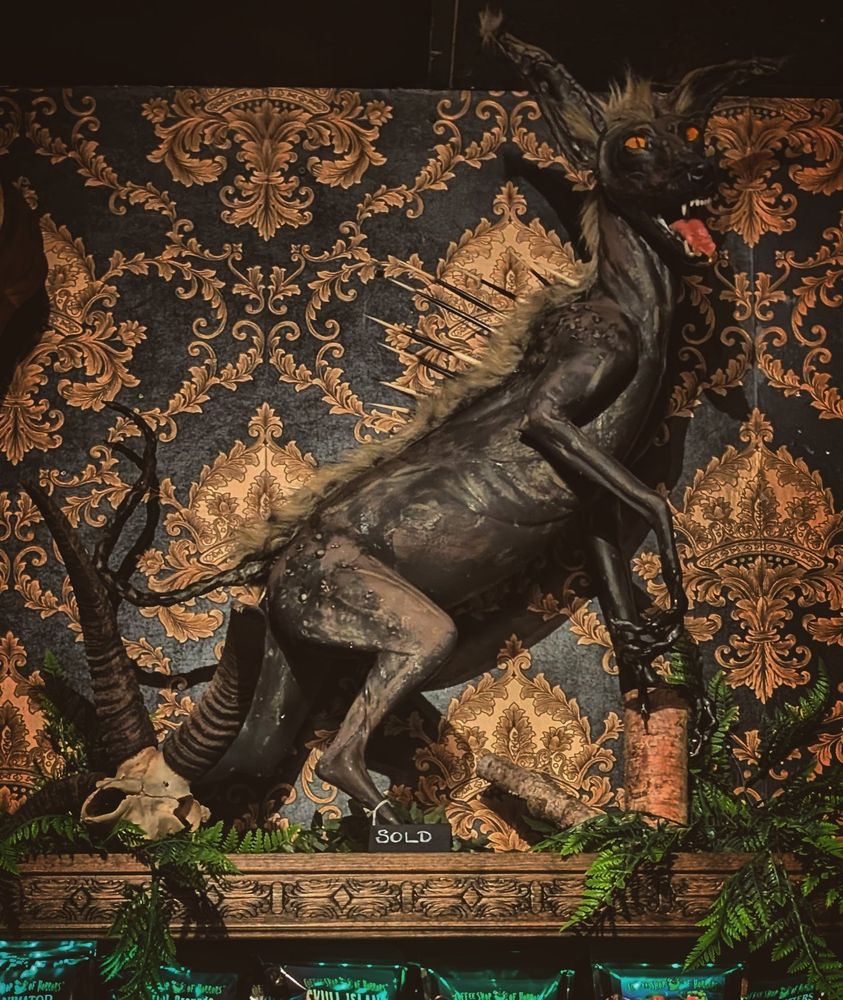 Faux taxidermy chupacabra (maybe) with a furless Kangaroo-Like body, thin bonytail, mohawk-like fur down the spine, porcupine quills along the spine, orange cat-like eyes, tall pointy ears, and a face like a canine. Against a black and gold damask background.