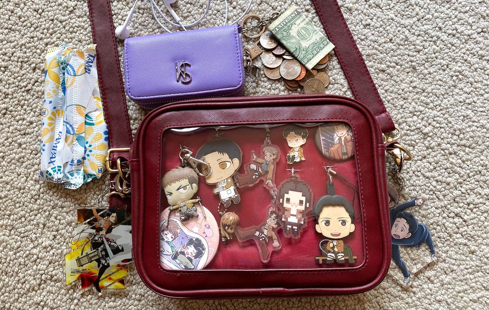 a burgundy ita bag full of pins and keychains of marco bodt (RIP) and jean kirstein from the anime Attack on Titan. this is in dedication to the jeanmarco ship that was popular on tumblr from like 2013-2015ish. the contents of the bag surround it and include a lavender wallet, some coins and cash, headphones, and tampons. i really don't carry much around i guess.