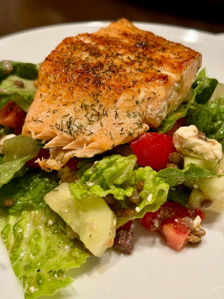 A salad of romaine, cucumber, tomato, feta, lentils, and dried dill, with a lemon Dijon vinaigrette, topped with a pan-seared salmon fillet. 