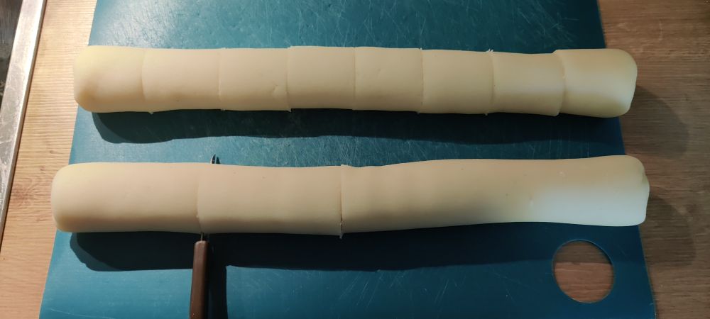 2 Rollen aus Kloßteig auf grünem Brettchen. Die obere gleichmäßig in 8 Teile geschnitten, die untere in der Mitte und in der Mitte der linken Hälfte zerschnitten. Im letzten Schnitt steckt noch ein Gemüsemesser mit braunem Griff.