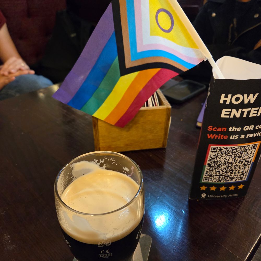 A pub table with a pint of Guinness and a pride progress flag.