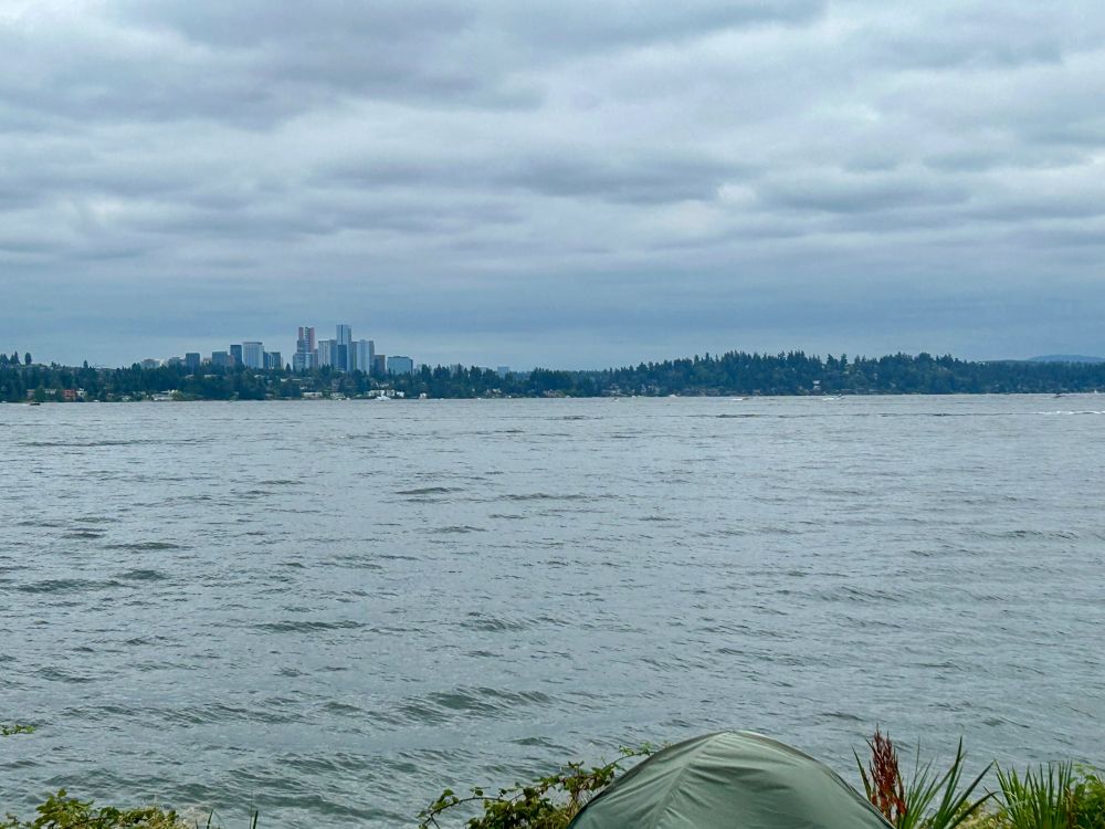 Bellevue city skyline with many skyscrapers is seen far across Lake Washington with choppy water and grey skies overhead. Across the lake the top of a tent and shrubbery is seen at the bottom. 