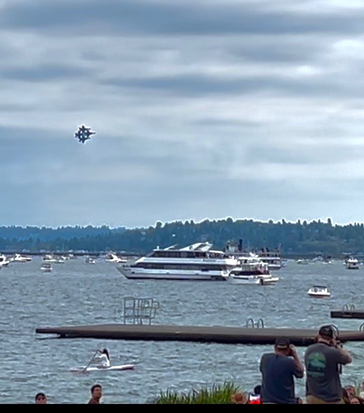 Blue Angels five jets are seen in diamond formation in the sky against an overcast sky above various sized boats in the water below, two guys are taking photos and others turning heads in the foreground at the bottom. A sole kayaker is seen close to shore.