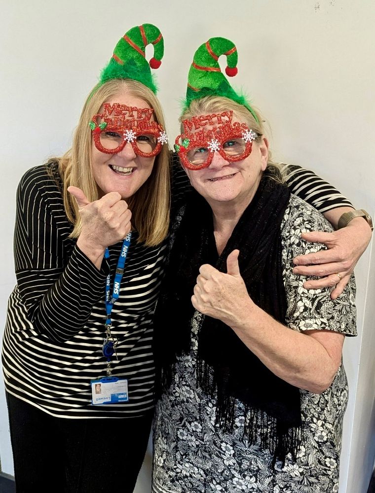 Two women wearing Merry Christmas glasses and elf hats, smiling with thumbs in the air