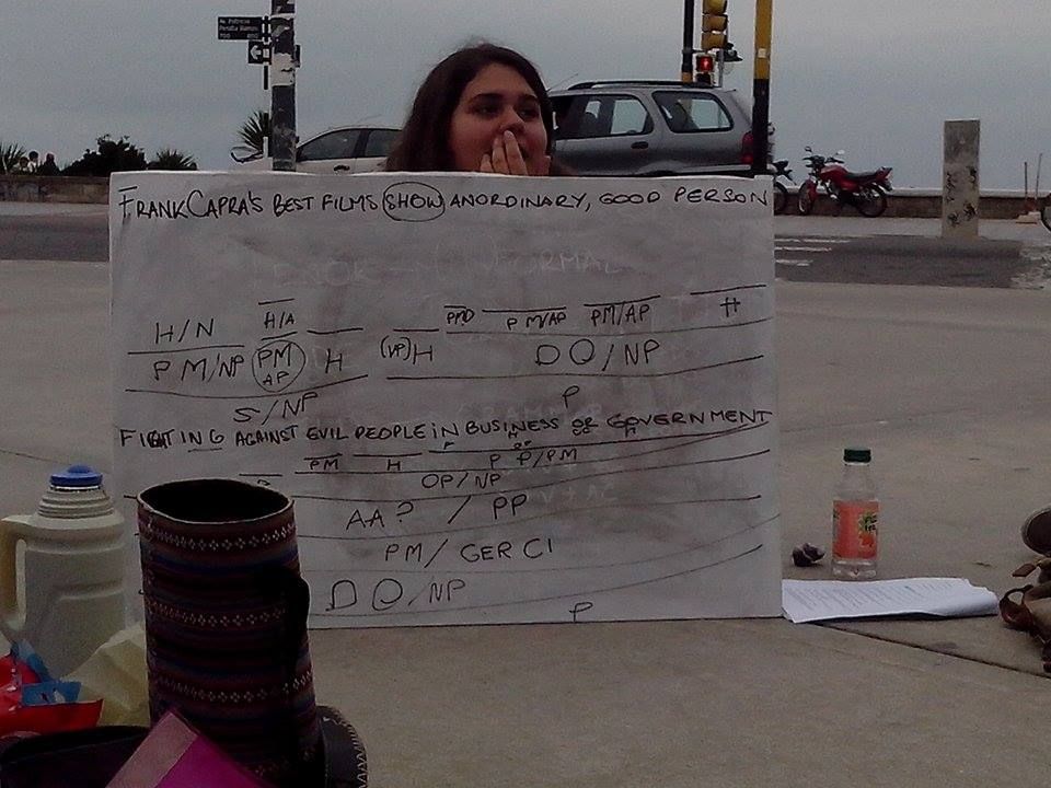 The author of this skeet is holding a whiteboard with a syntactic analysis of a sentence. She is sitting on the floor and the whiteboard is covering her whole body. One can see a thermos and a mate set from the students who were sitting around in a circle. The picture was taken at Plaza España, Mar del Plata, Argentina, which is right by the Atlantic coast, on a cloudy day.