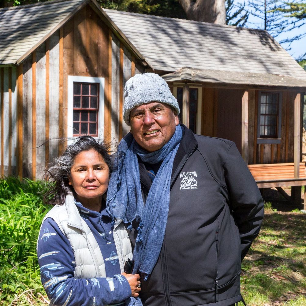 A woman and a man in jackets and scarf standing close together in front of an old cabin under a tree.