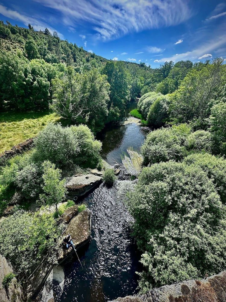 A river as viewed from above within a lush valley filled with trees and large rocks below and open sky.