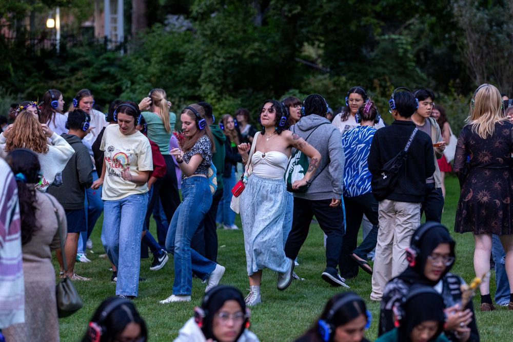In a verdant garden setting, a crowd of diverse students gather, dancing to the beat of music playing in their glowing headphones at a silent disco.