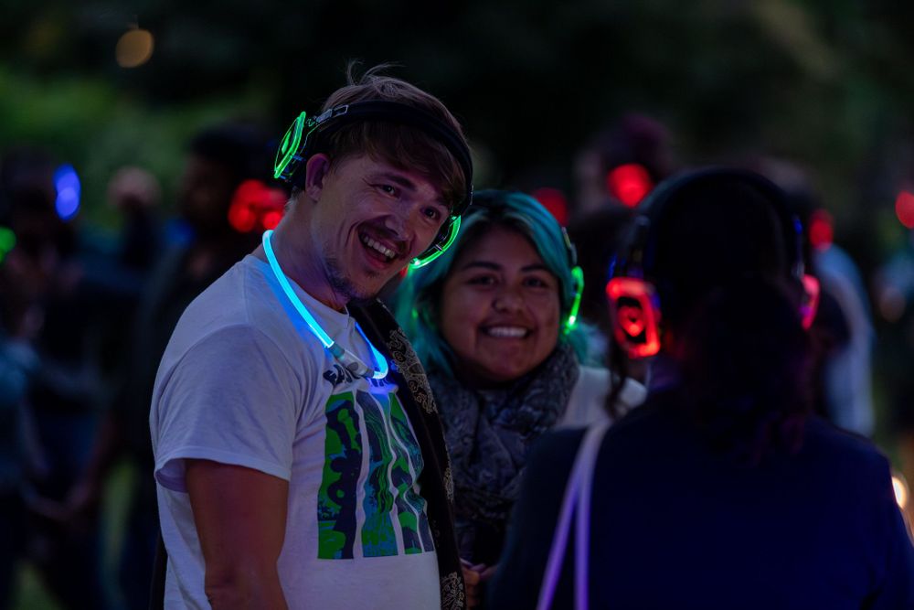 Two MSU students wearing glowing headphones outdoors smile and laugh, others wearing headphones visible but blurry in the dark of evening.