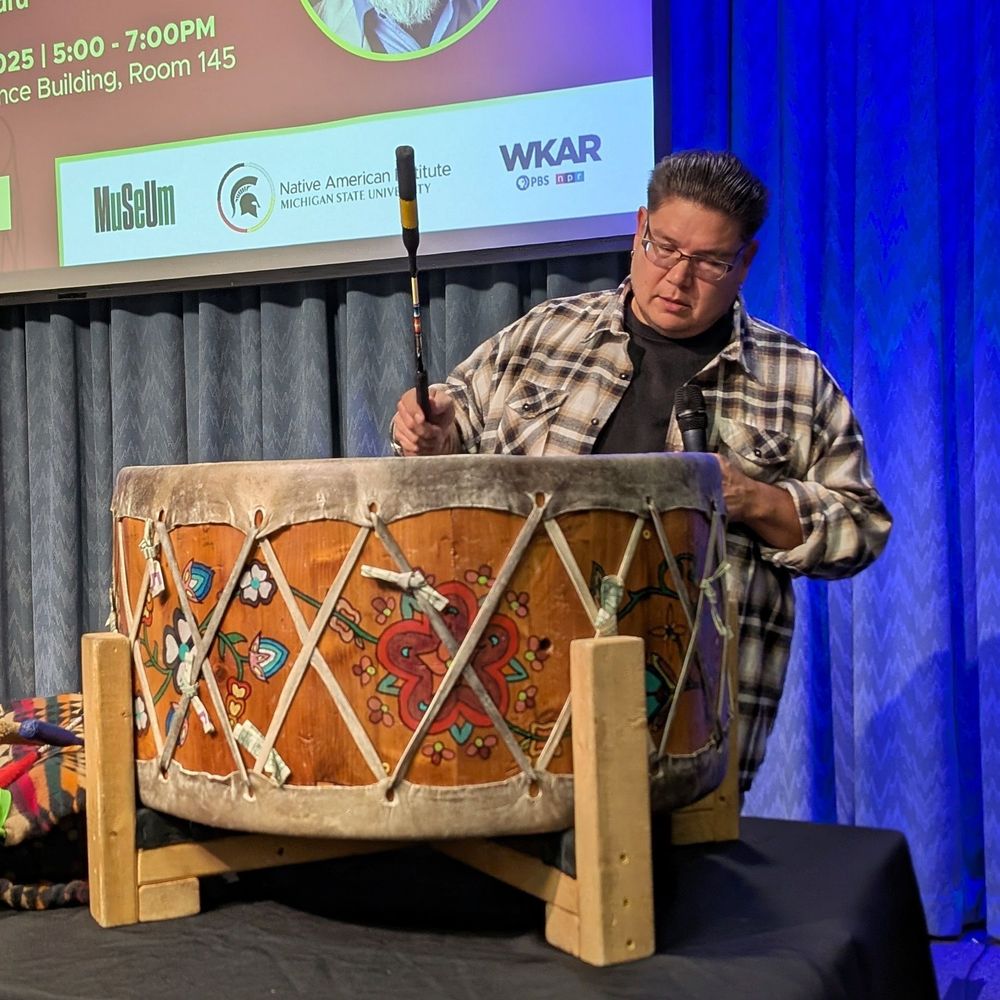 A Native American man of the of The Saginaw Chippewa Indian Tribe of Michigan stands in front of a large wooden drum. He wears a tan and black plaid shirt and black glasses, beating a large drum with a black drumstick. Around the surface of the drum are illustrations of flowers and wide lacing the connects the hide surface on the top and bottom.