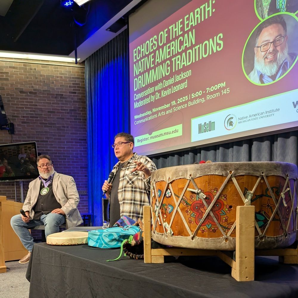 A wide view of a stage, focusing on a large decorated wood and hide drum resting on wooden supports on a table covered in a black cloth. Behind the drum, two men holding microphones stand and discuss the items on the table. The moderator wears a tan blazer and jeans while the guest on the right wears a tan and black plaid shirt, glasses, and jeans.