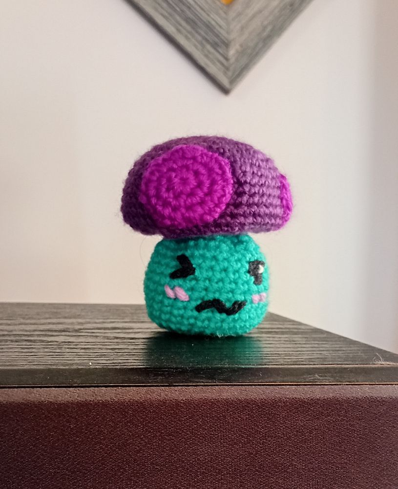 A crocheted mushroom with a teal stalk and purple top with large pink spots. It has an embroidered face with one closed eye, one open eye, and a squiggly mouth. It's blushing.