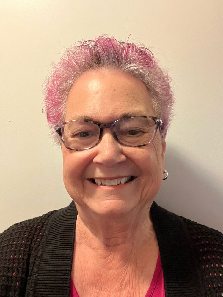 Selfie with glasses and purple and gray hair, wearing a black sweater over a pink shirt.
