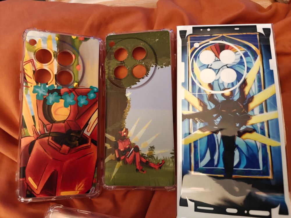 3 phone cases and sticker wrap for the oneplus 12 are layed out on a blanket. 2 cases feature V2 from uktrakill, one wearing a flower crown, the other of V2 sittung under a tree. The 3rd phone case is transparent. The sticker wrap depicts V2 in shadow, in front of stained glass from 1-4 of the game where you first meet them.