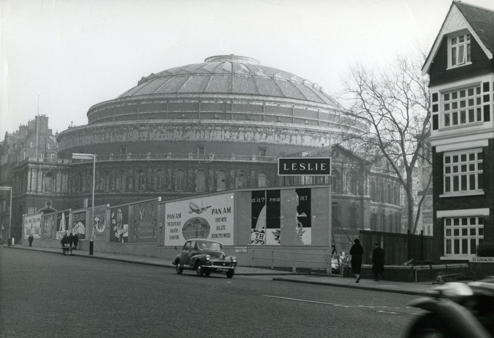 Black and white photo showing the site of RCA Kensington before it was built with hoardings and advertisement around the building site, cars on the road in front and the Royal Albert Hall in the background.