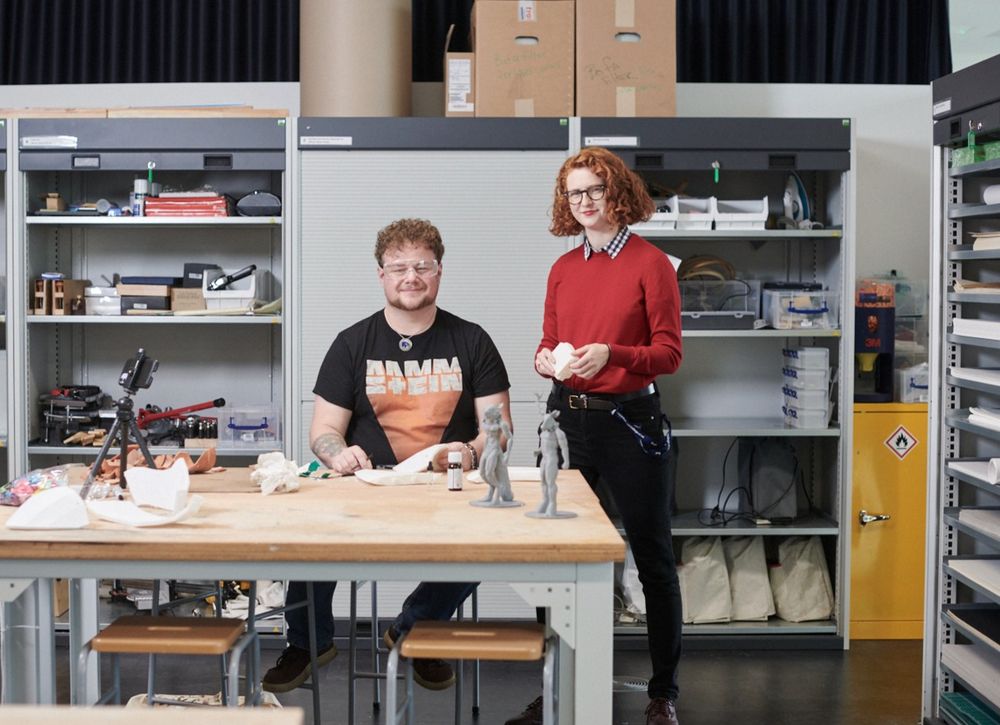 Two individuals working in a workshop, one seated and one standing, surrounded by tools and prototype models on a worktable.