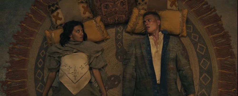Egwene and Rand lying on the ground apart from each other. Both of them looking at each other with confusion. Egwene is wearing a grey shawl (possibly?) and a white shirt. Whilst Rand is wearing a battered up jacket with blood stains and white shirt underneath.
