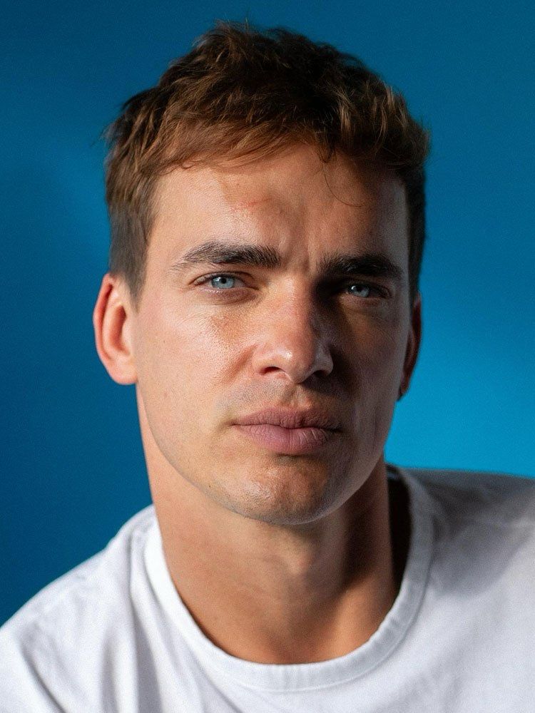 Headshot of actor Luke Fetherston looking into the camera. He’s wearing a white t-shirt and has striking blue eyes. He has short fluffy red hair. 