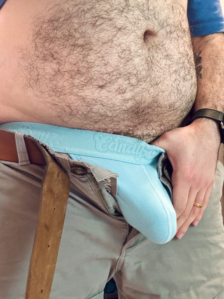 Hairy belly man with unzipped shorts, his jockstrap bulges past the zipper. Hand by bulge with thumb tucked into the blue jockstrap