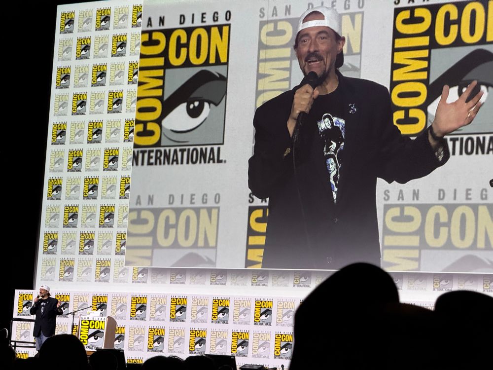Kevin Smith on stage with a video behind him of himself in Hall H
