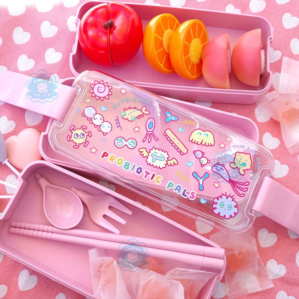 Photograph of a stackable bento lunchbox designed by marinewaltz. The lid has a whimsical design, with colourful text of "PROBIOTIC PALS" surrounded by cute little creatures who are happily floating around. There are matching utensils and prop fruits sitting inside the bento layers, and decorative hearts and a picnic blanket underneath.