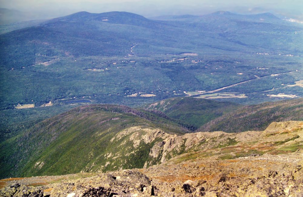 Beneath a bright sunny sky, hazy white along the horizons, which are all that is visible of the sky in this photo. We are standing atop a lofty mountain looking down a long slope covered with gray granite rock and occasional low green and golden brown ground cover. There is a sprawling valley far below, mostly wooded, with several visible roads. A few large green fields break up the forests, mostly on or near the roads. We can also see one large building and a scattering of houses. A line of wooded hills runs down the middle of the valley, which soon disappears into the haze along the horizon.
