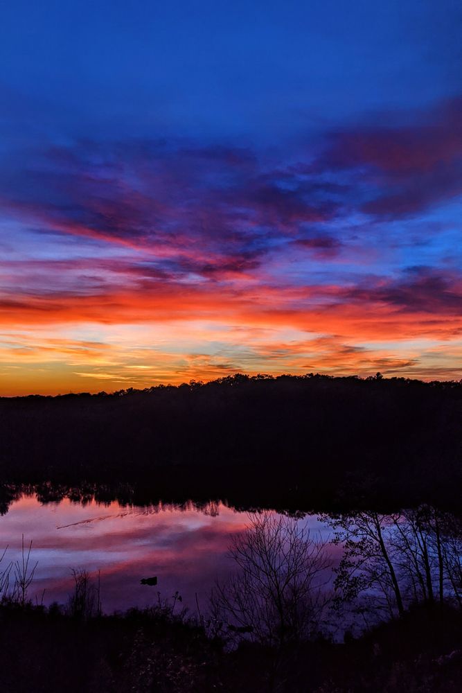 A late post sunset shot looking down from a ridge crest. The low hills and forest surrounding a small lake below are almost completely silhouetted. The calm lake surface mirrors the shy above it, in which streaky clouds are being tinted pink, orange and gold shades by the recently set sun. The clouds' shadows are a deep purple. Above it all, a deep blue twilight descends upon the scene.