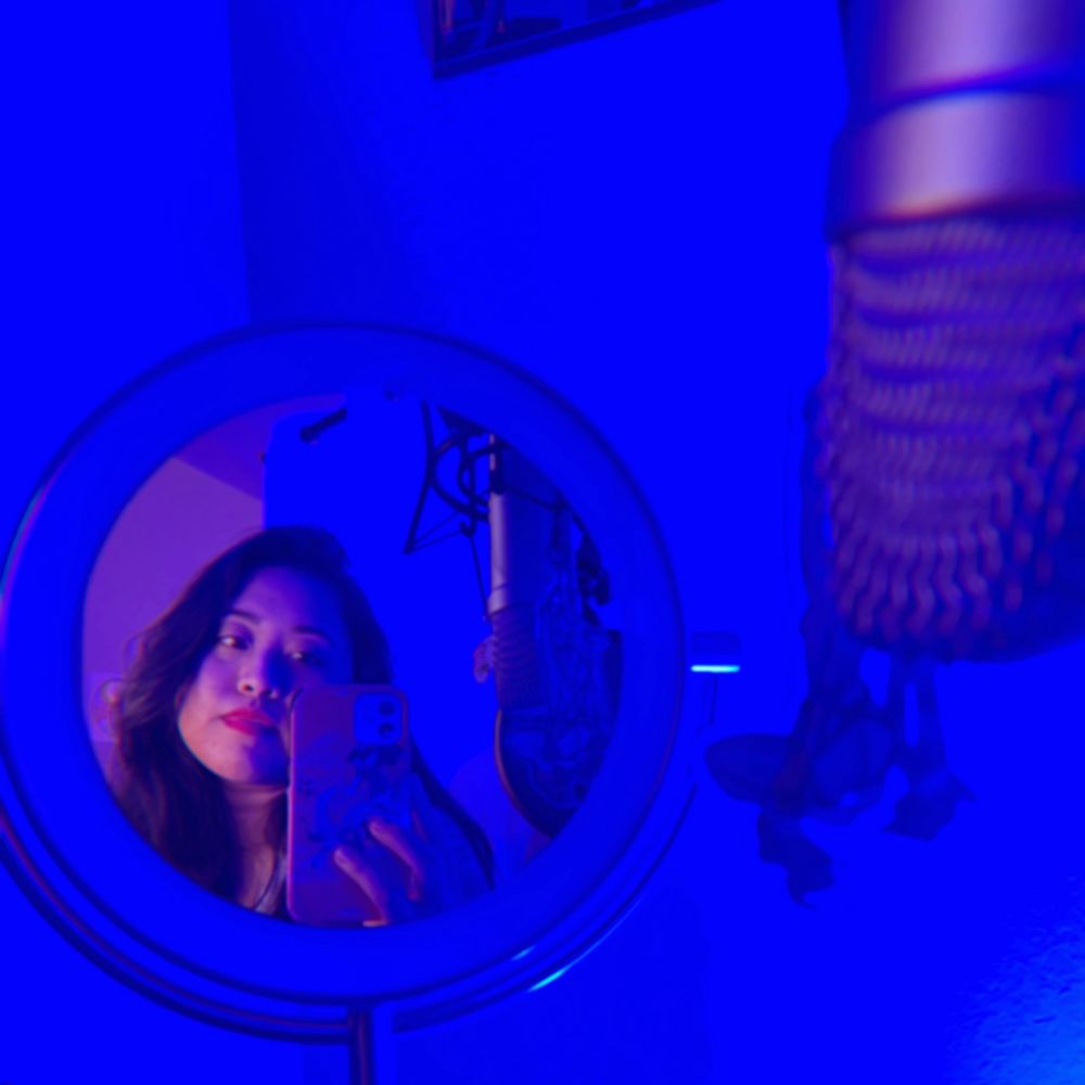 Woman with black hair and red lipstick takes a photo of herself looking in a small circular standing mirror on her desk with her smartphone. In the mirror and on the right side of the image is a microphone in a shock mount on a boom arm. The lighting is dark blue but in the mirror, there is some warm reddish purple lighting on her face