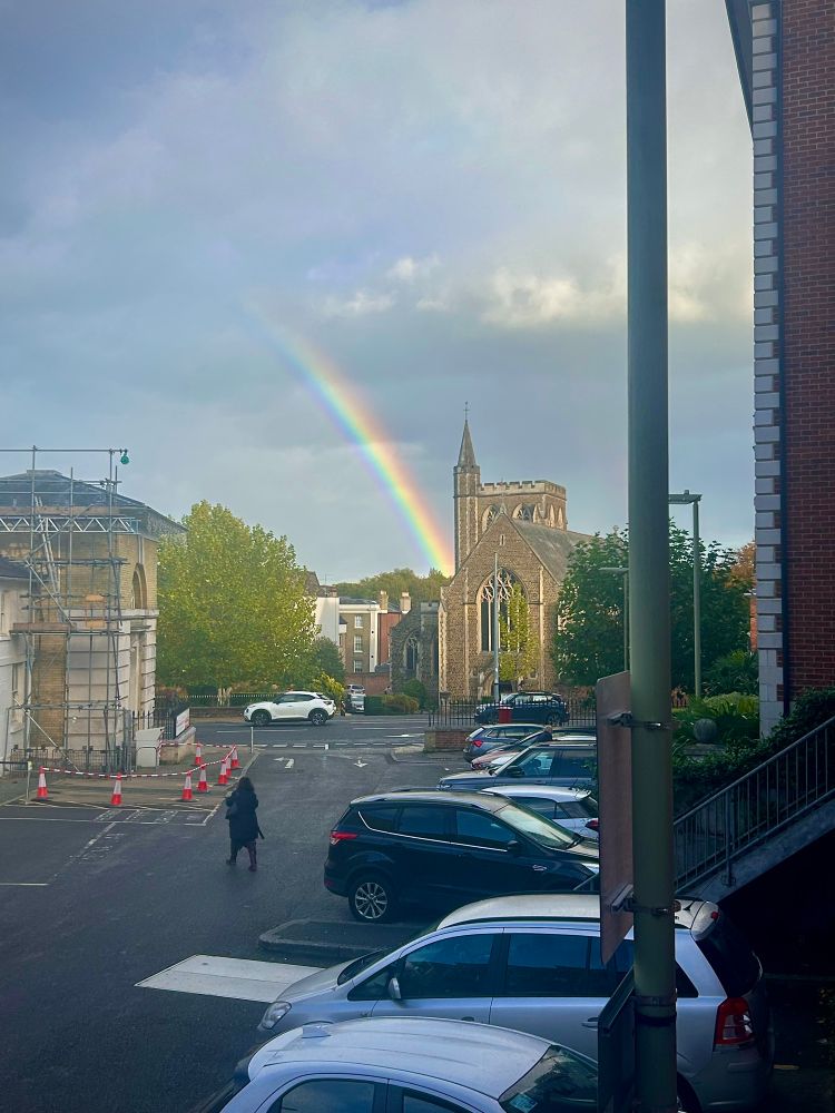 A rainbow, looks like it’s coming out of a church. Surrounded by grey skies 