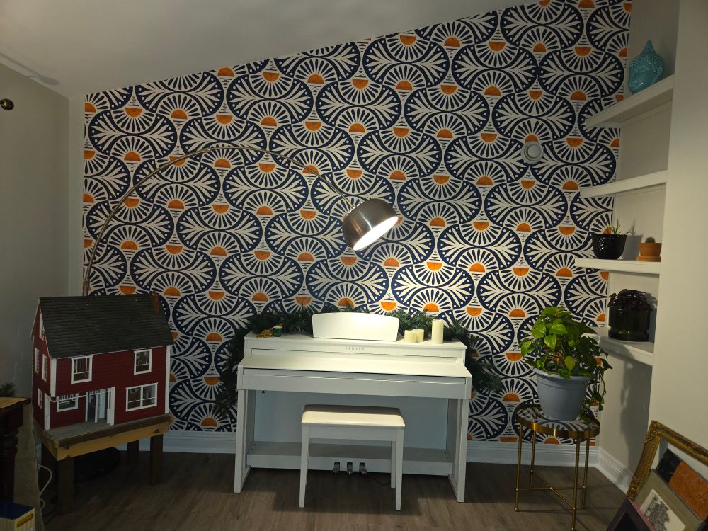 Room with wallpaper on one tall wall: large navy blue/ white/sunset orange scale type pattern. Looks like a sunset on the water with navy blue sun bursts. Infront of wallpaper in the center is a white upright piano, to the left of that is a red dollhouse and to the right of the piano is a plant on a gold end table. Also there's a spaceship look lamp that arches over the dollhouse and kinda floats above the piano