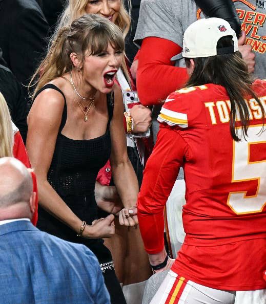 In this high-energy moment, Taylor Swift is front and center, leaning forward with her mouth wide open in an excited yell, her face lit with pure adrenaline and joy. She’s wearing a sleeveless, form-fitting black top and layered gold necklaces, with her hair in a loose ponytail and bangs framing her face. Her fists are clenched tightly at her sides in a celebratory gesture, and she’s angled toward a Kansas City Chiefs player in a bright red jersey with gold-and-white trim. The player, wearing the number 5 and the name “Townsend” on his back, has long brown hair flowing out from under a backward white cap. The background is packed with people, many in Chiefs colors, and the air is electric with post-game victory energy. This is the kind of unfiltered, joyous reaction that feels like a sports highlight in itself, radiating pure fandom and shared triumph.