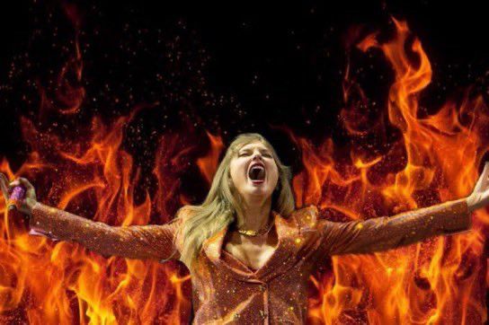 The image shows Taylor Swift, during a moment from her Eras Tour, edited to appear as if she is surrounded by towering flames. She is wearing a sparkling orange suit jacket with matching pants, shimmering under the stage lights. Her head is tilted slightly back, mouth open wide in an expression that looks like she’s singing powerfully or shouting triumphantly. Her arms are extended outward, as if embracing the crowd or declaring victory, with her palms open. The background has been digitally altered to depict vivid, roaring fire in shades of bright orange, red, and yellow, with tongues of flame curling upwards against a black backdrop, creating a dramatic and intense visual effect. The flames frame her figure entirely, giving the impression of an unstoppable, commanding presence, almost like a fiery goddess on stage. The scene conveys energy, dominance, and passion, with Taylor at the center of a blaze that mirrors her performance intensity.