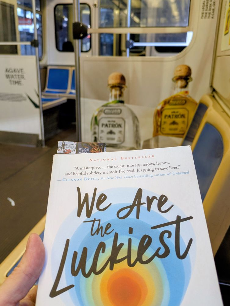 In a train car, cover of the book "We are the Luckiest, the Surprising Magic of a Sober Life" with posters of Patron tequila in the background.
